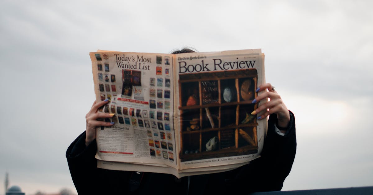 Individual holding a book review newspaper with colorful nails against a cloudy sky backdrop.